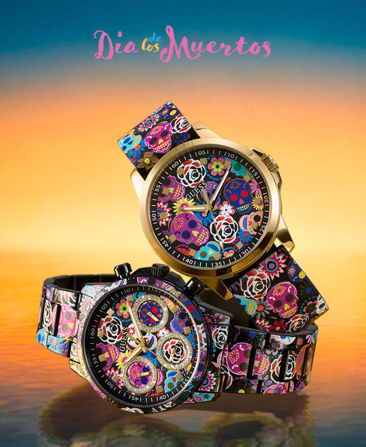 Two gold-tone Dia de los Muertos themed watch with colorful sugar skull and floral patterns on the straps and dial, set against a sunset gradient background with the text 'Dia de los Muertos' at the top.
