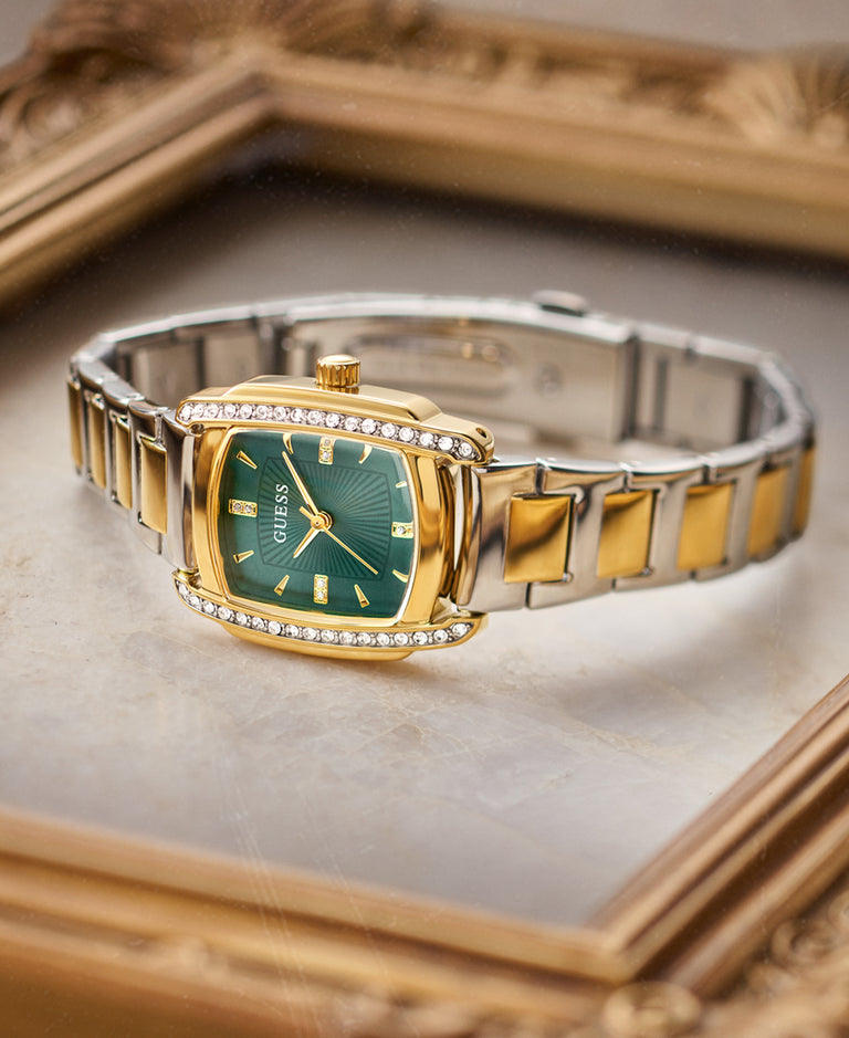 A Guess women's watch with a gold-tone case, green dial, and a two-tone silver and gold link bracelet, resting on a light marble surface framed by an ornate gold picture frame.