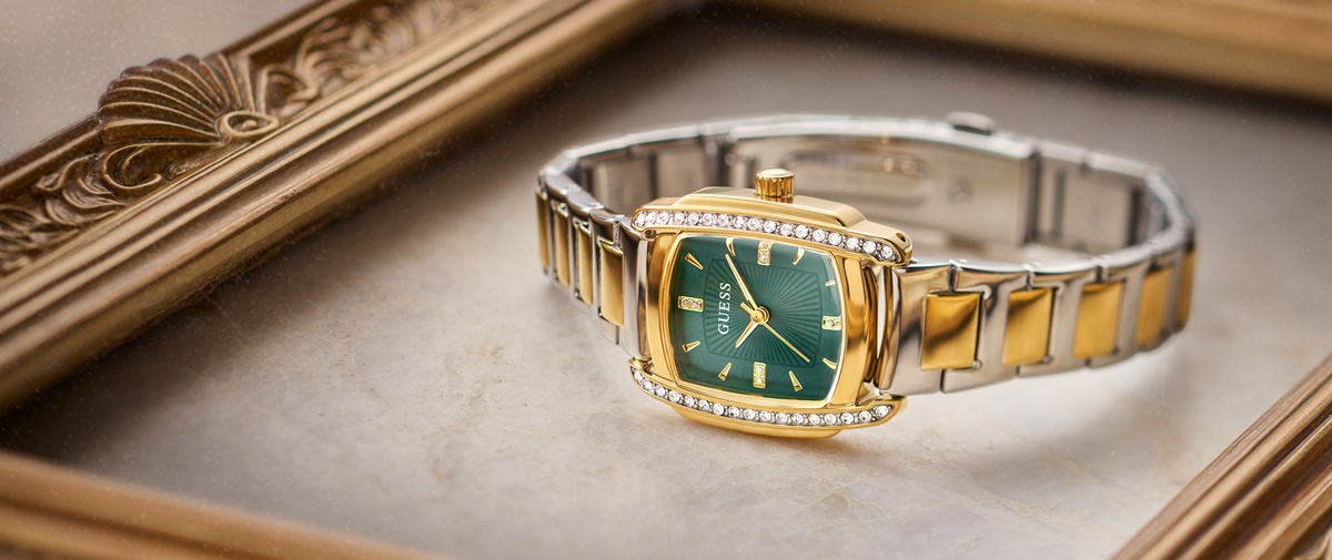 A luxury wristwatch with a gold and silver two-tone metal bracelet, a green dial, and a bezel set with small diamonds, resting on a beige marble surface next to an ornate gold picture frame.