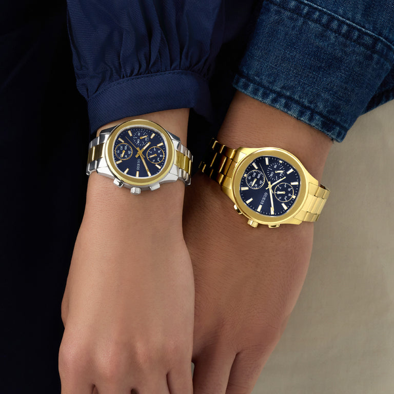 Two wristwatches, one silver and one gold, on a person's wrist against a neutral background.