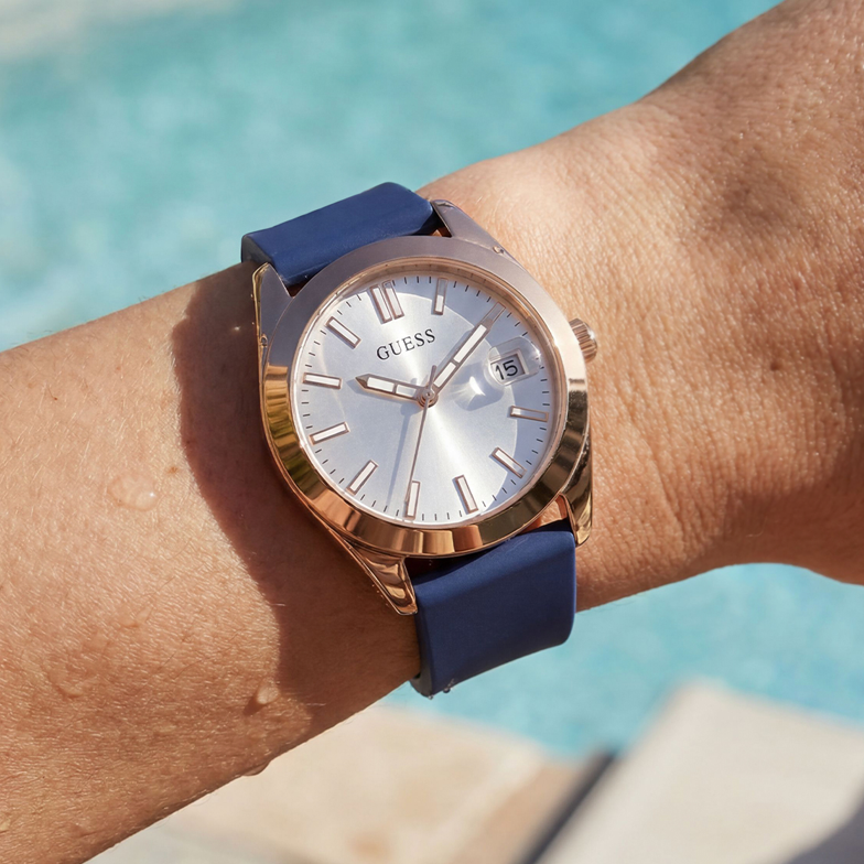 Wrist wearing a Guess watch with a blue strap and rose gold case by a pool.