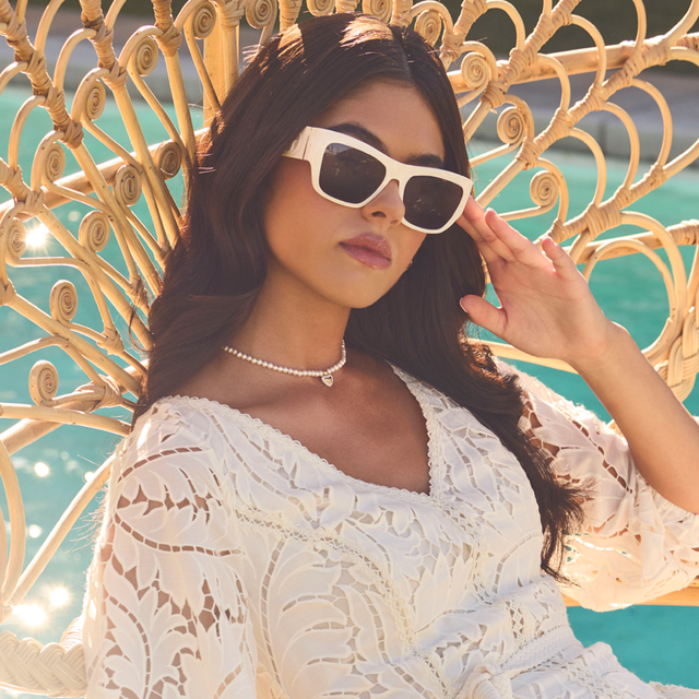 Woman in a white lace dress and sunglasses standing in front of a decorative metal screen with a blue background.