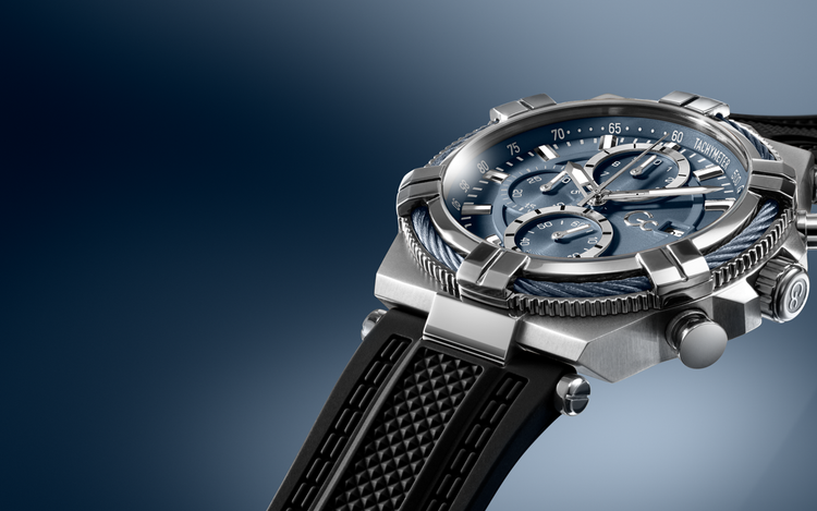 A close-up studio shot of a luxury chronograph watch with a silver-toned metal case, blue dial, and black textured strap against a dark blue gradient background.
