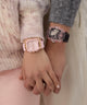 Two wrists wearing pink watches held together, with a neutral background.