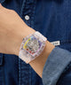 Wrist wearing a transparent GUESS Mens Pride Limited Edition Clear Analog Watch with colorful inner design, against a denim background.