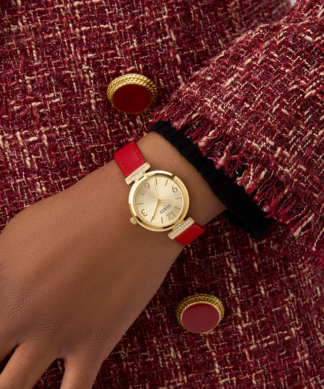 GUESS Ladies Red Gold Tone Analog Watch with a red strap on a wrist against a textured maroon background