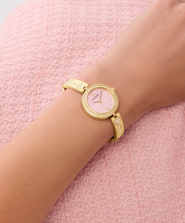 Gold GUESS Ladies Gold Tone Analog Watch on a wrist with a pink textured background