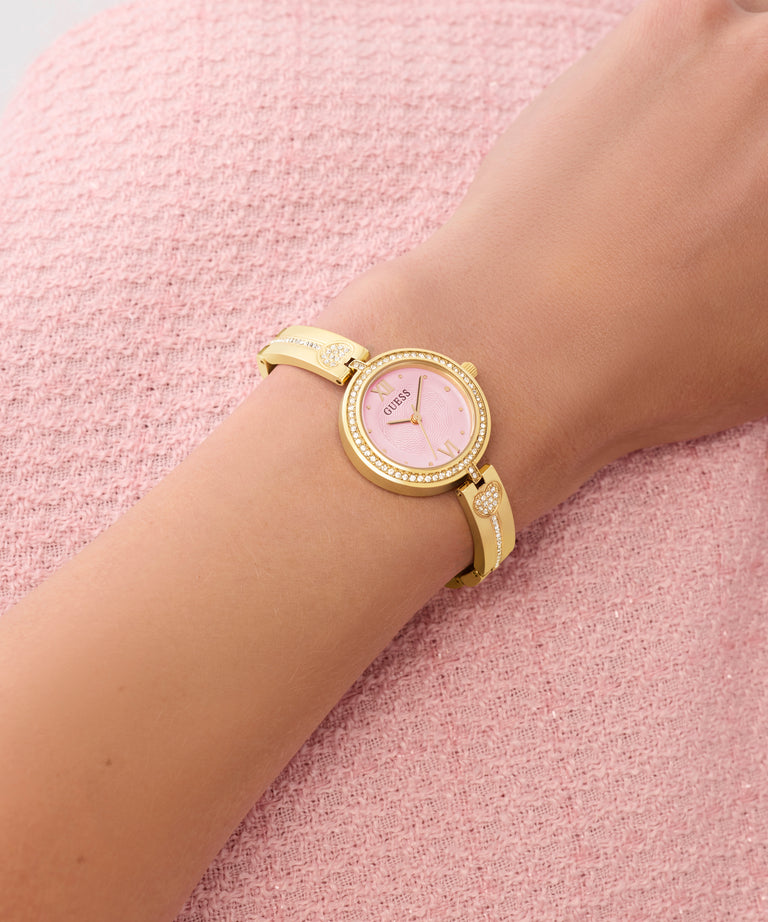 Gold GUESS Ladies Gold Tone Analog Watch on a wrist with a pink textured background