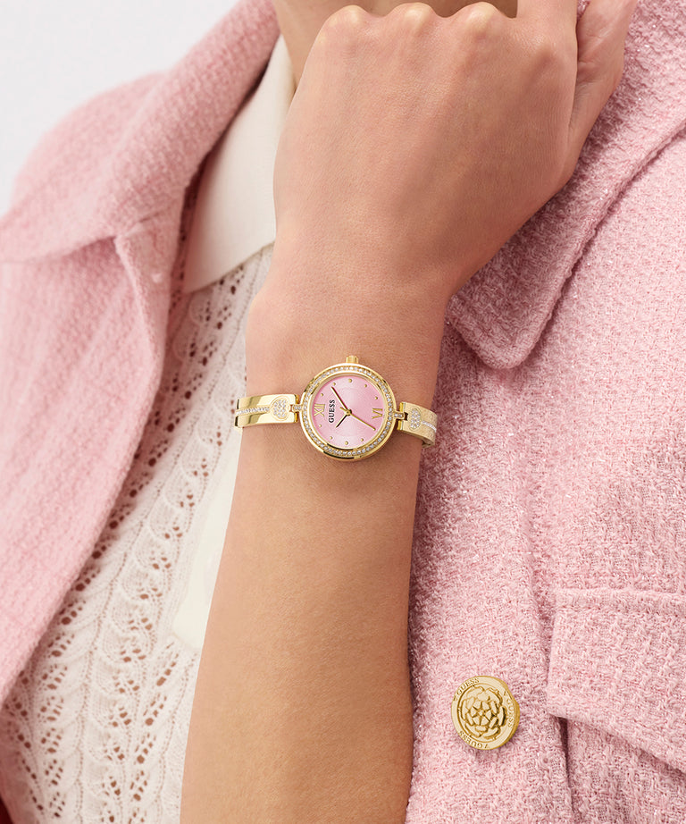Gold watch on a wrist with a pink face, worn against a pink textured background.