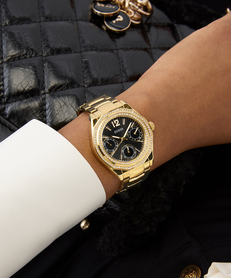 GUESS Ladies Gold Tone Multi-function Watch on a wrist with a black quilted bag in the background