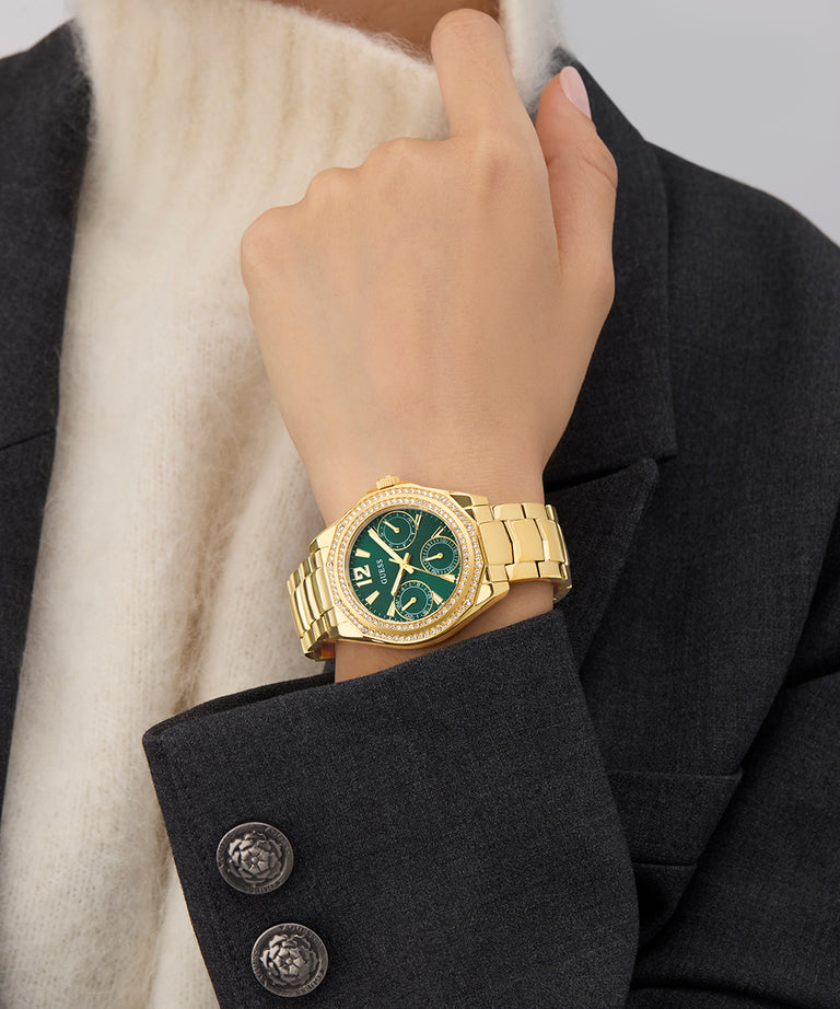 GUESS Ladies Gold Tone Multi-function Watch with green face on a person's wrist, wearing a dark coat and white scarf.