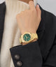 GUESS Ladies Gold Tone Multi-function Watch with green face on a person's wrist, wearing a dark coat and white scarf.