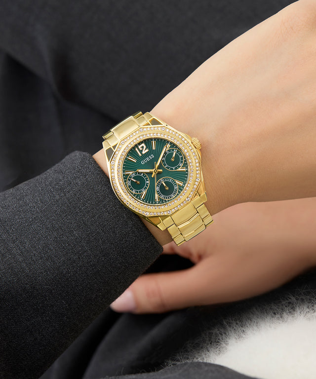 GUESS Ladies Gold Tone Multi-function Watch with a green face on a person's wrist, wearing a dark suit.