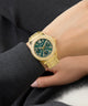 GUESS Ladies Gold Tone Multi-function Watch with a green face on a person's wrist, wearing a dark suit.