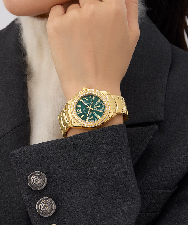 GUESS Ladies Gold Tone Multi-function Watch with a green face on a person's wrist, wearing a dark suit.
