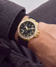 A gold-tone GUESS watch with a black dial and a dark blue or black textured strap, worn on a wrist with a gray knit sweater cuff visible.