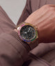 A close-up of a wrist wearing a luxury watch with a rose gold-toned case, a black dial, and a bezel encrusted with multicolored gemstones in pink, yellow, green, and blue, paired with a brown textured strap.