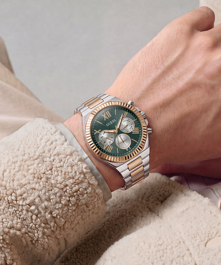A close-up of a wrist wearing a Guess watch with a green dial, gold and silver two-tone metal case, and matching metal bracelet, styled with a beige fleece jacket.