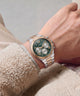 A close-up of a wrist wearing a Guess watch with a green dial, gold and silver two-tone metal case, and matching metal bracelet, styled with a beige fleece jacket.