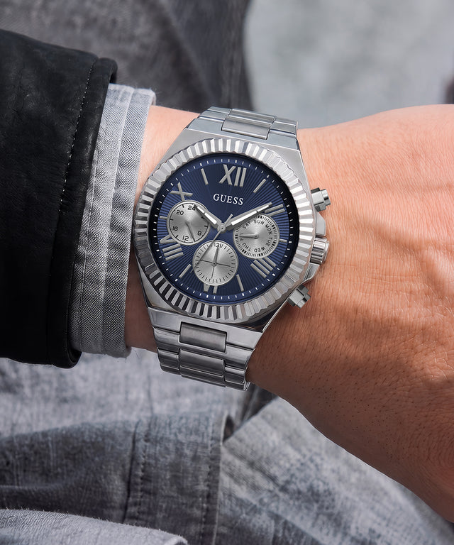 A silver stainless steel wristwatch with a blue dial and Roman numeral markers, shown worn on a person's wrist. The watch has a fluted bezel, a three-subdial chronograph layout, and a matching steel link bracelet.