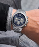 A silver stainless steel wristwatch with a blue dial and Roman numeral markers, shown worn on a person's wrist. The watch has a fluted bezel, a three-subdial chronograph layout, and a matching steel link bracelet.