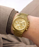A gold-tone Guess analog watch with a matching metal link bracelet, worn on a person's wrist. The watch features a fluted bezel, Roman numeral hour markers, and three sub-dials on a gold dial.