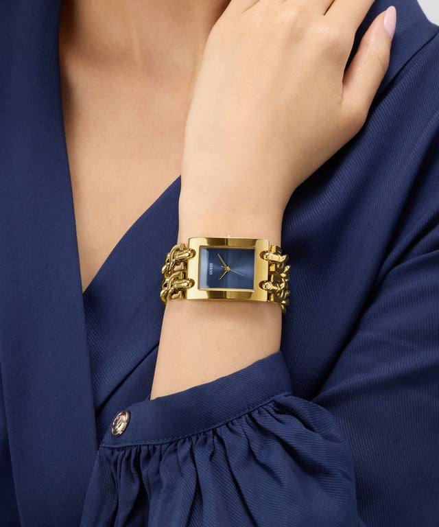Gold GUESS Ladies Mod Heavy Metal Gold Tone Analog Watch on a wrist with a blue dress