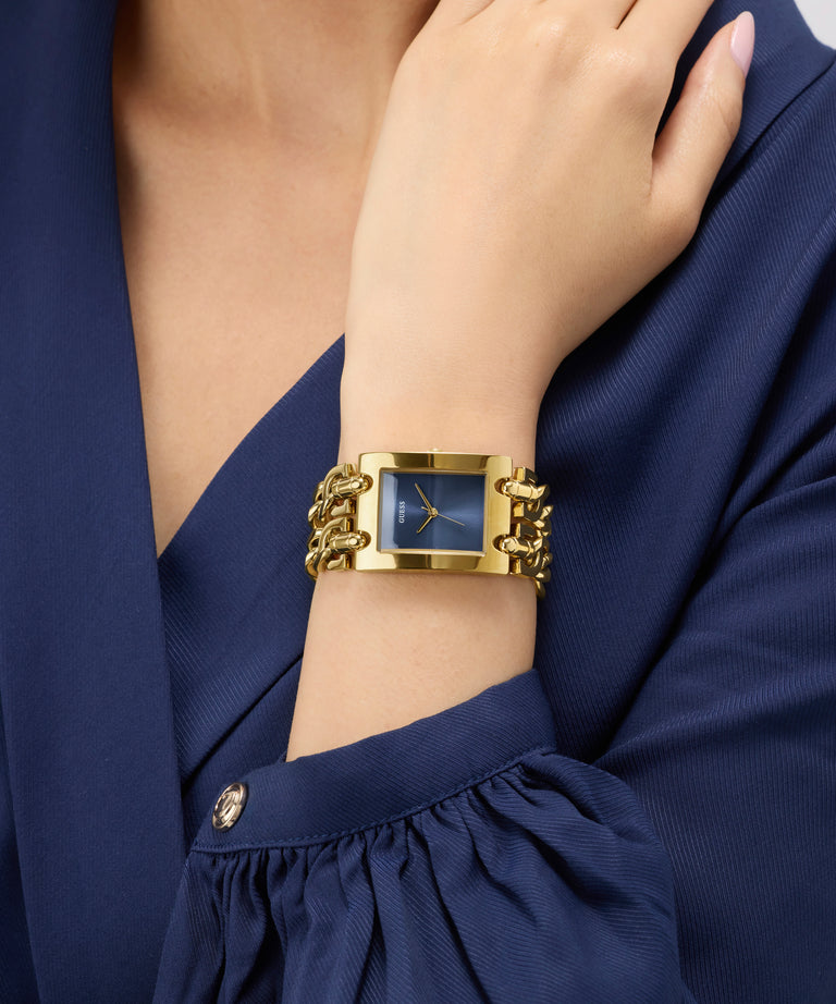 Gold GUESS Ladies Mod Heavy Metal Gold Tone Analog Watch on a wrist with a blue dress