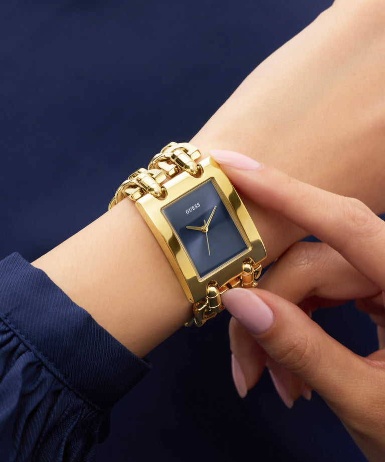 Gold rectangular GUESS Ladies Mod Heavy Metal Gold Tone Analog Watch on a wrist with a dark blue background