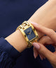 Gold rectangular GUESS Ladies Mod Heavy Metal Gold Tone Analog Watch on a wrist with a dark blue background
