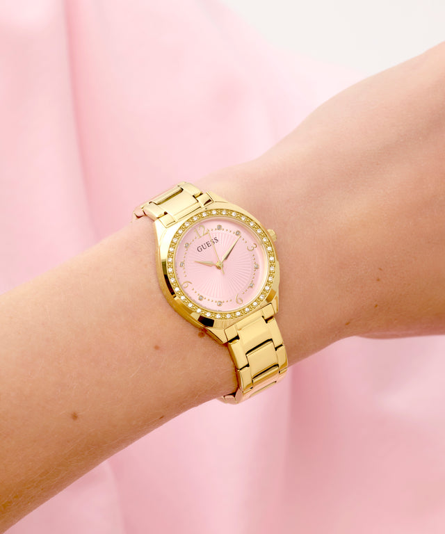 Gold GUESS Ladies Gold Tone Analog Watch on a wrist with a pink background