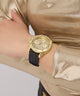 GUESS Ladies Black Gold Tone Multi-function Watch with a black strap on a wrist, wearing a gold outfit.