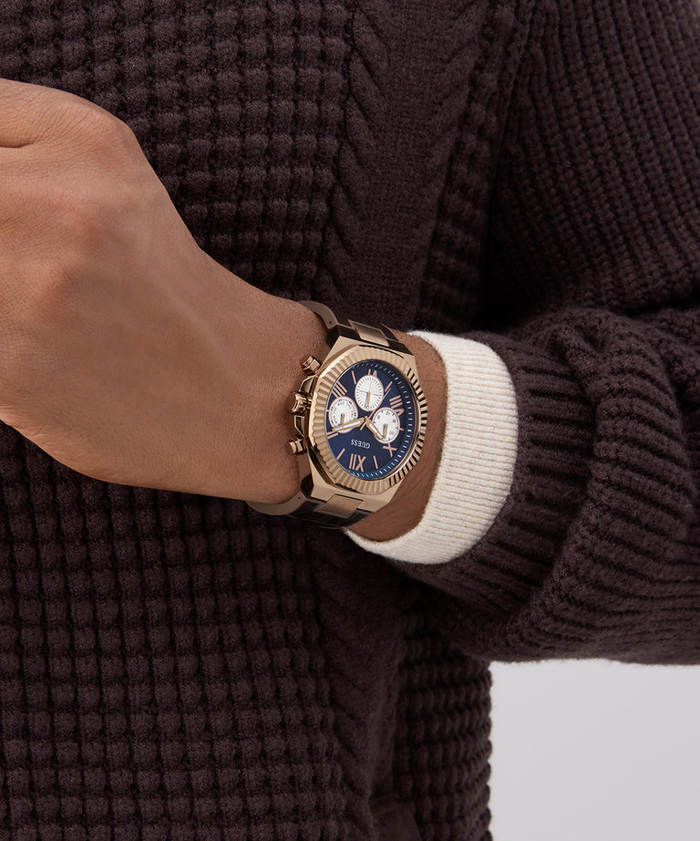 Close-up of a hand wearing a gold watch with a blue face on a brown textured sweater.