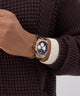 Close-up of a hand wearing a gold watch with a blue face on a brown textured sweater.