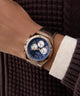GUESS Mens Brown Coffee Multi-function Watch with blue face and rose gold case on a wrist, wearing a dark textured sweater.