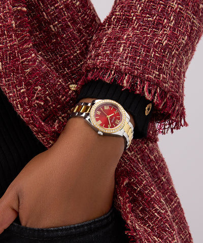 Hand wearing a GUESS Ladies 2-Tone Analog Watch with a red face and gold accents, wrapped in a textured red scarf.
