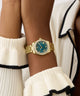 Gold watch with a teal face on a wrist, wearing a white and black outfit.