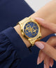 Gold GUESS Ladies Gold Tone Analog Watch with blue face on a wrist, against a dark background