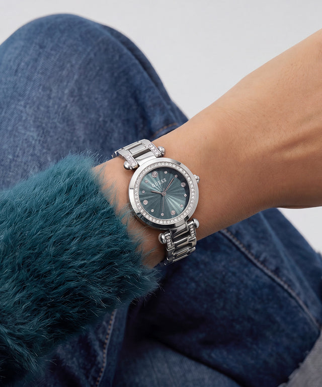 A silver-tone stainless steel watch with a textured dark gray dial and a bezel set with small clear stones, shown on a person's wrist wearing blue jeans and a teal fuzzy sleeve.
