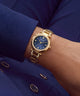 A gold-tone guess watch with a blue sunburst dial and crystal-encrusted bezel, shown worn on a wrist against a navy blue blazer background.