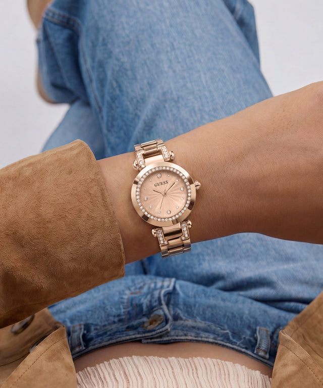 A close-up of a woman's wrist wearing a rose gold GUESS watch with a crystal-studded bezel and matching metal link bracelet, styled with blue denim shorts and a brown suede jacket.