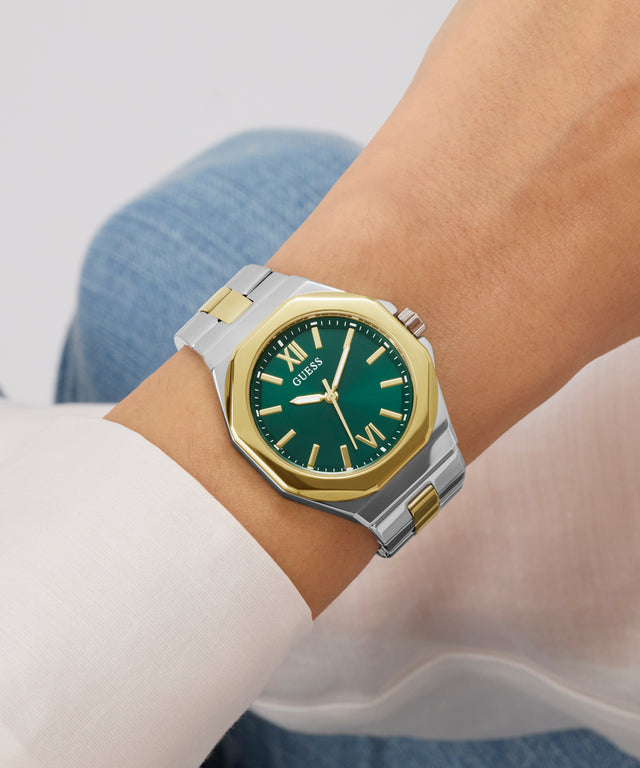 Wrist wearing a green and gold watch with a white shirt and blue jeans.