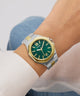 Wrist wearing a green and gold watch with a white shirt and blue jeans.