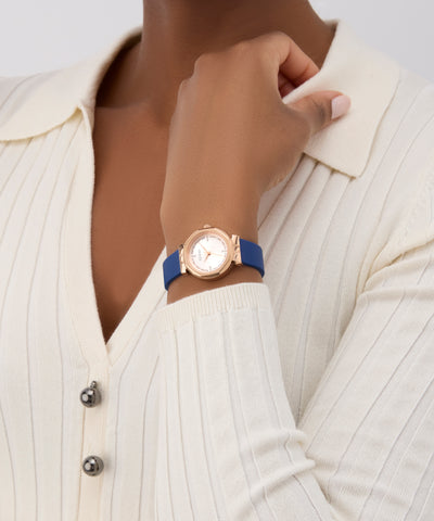 Person wearing a GUESS Ladies Blue Rose Gold Tone Analog Watch with a blue strap and gold case on a white background