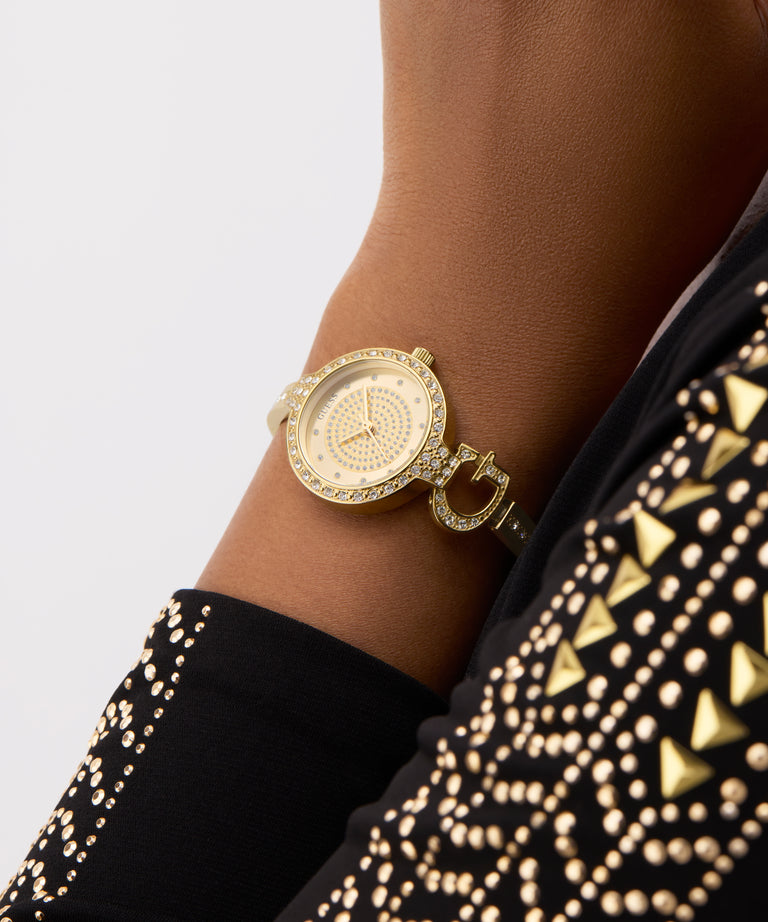 GUESS Ladies Gold Tone Analog Watch on a wrist with a black and gold patterned sleeve against a light gray background