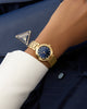 Gold watch on a wrist with a blue dress shirt and navy suit jacket, featuring a Guess logo. GW0930L4