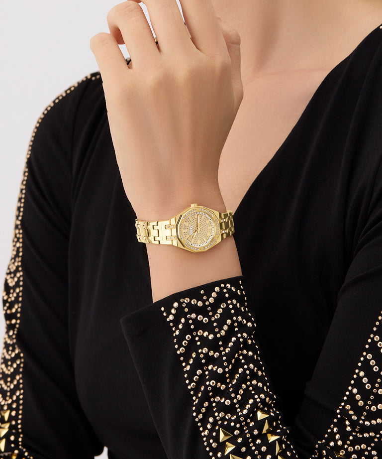 Gold GUESS Ladies Gold Tone Analog Watch on a wrist with a black garment featuring gold embellishments.