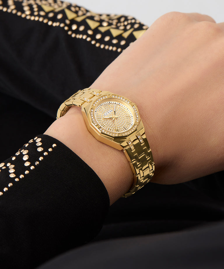 Gold GUESS Ladies Gold Tone Analog Watch on a person's wrist with a black garment featuring gold embroidery in the background