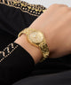 Gold GUESS Ladies Gold Tone Analog Watch on a person's wrist with a black garment featuring gold embroidery in the background