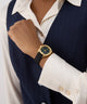 Hand wearing a GUESS Ladies Black Gold Tone Analog Watch on a person in a suit with a white shirt.